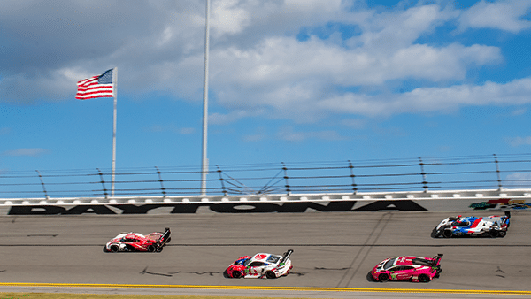 Cars in IMSA race on bank of Daytona Speedway track with blue sky and clouds above